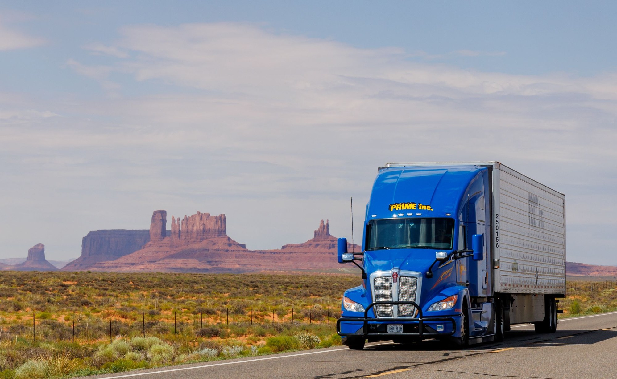 A blue American semi-truck with an enclosed silver trailer travels along a desert highway beneath Monument Valley mesas.