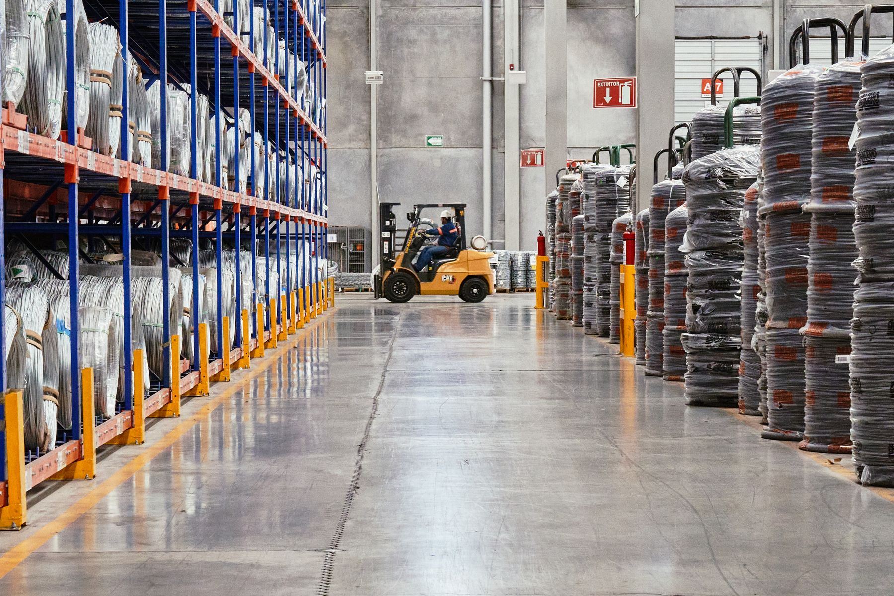 A clean industrial warehouse aisle has tall pallet racks, wrapped coils and a yellow forklift in the background.
